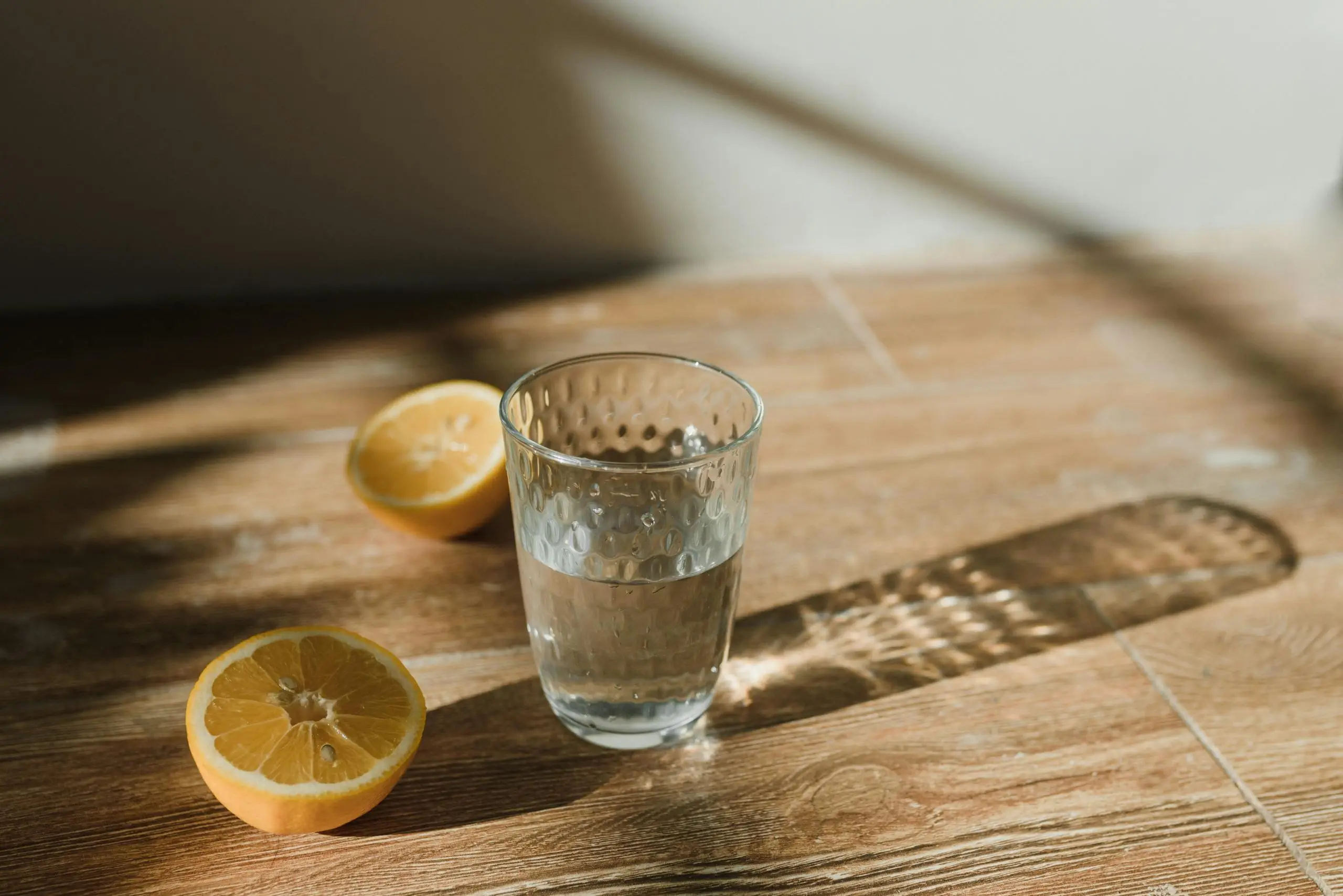 a glass of water and two lemon halves are placed on a wooden surface with sunlight casting shadows Clear glass of water with sliced lemons on a wooden surface in natural sunlight.